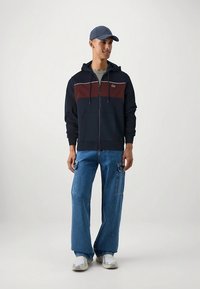 Tengerészkék cipzáros kapucnis pulóver bordó mellrész betéttel, alatta szürke póló, széles kék farmernadrággal és fehér sportcipővel párosítva. Laza sportos öltözék.