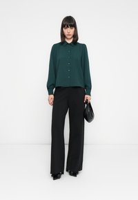 Mørkegrønn bluse med knappeåpning og krage, kombinert med løse svarte bukser. Outfiten er komplettert med en svart håndveske.