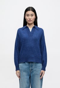 Blauer Strickpullover mit V-Ausschnitt und Kragen, lockere Ärmel und eine entspannte Passform. Kombiniert mit hellblauen Jeans, weicher Textur.