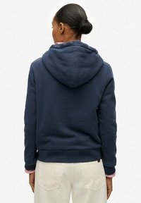 Mujer con el cabello recogido en un moño, vestida con una sudadera con capucha azul marino y pantalones beige claros, vista de espaldas sobre un fondo blanco.