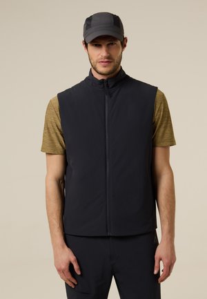 Hombre que lleva un chaleco sin mangas negro con cremallera, camiseta de manga corta mostaza, pantalones negros y una gorra negra, de pie frente a un fondo liso.