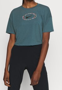 Persona che indossa una maglietta cropped color teal con logo Nike bordato di fiori e leggings neri a vita alta, in piedi su uno sfondo semplice.