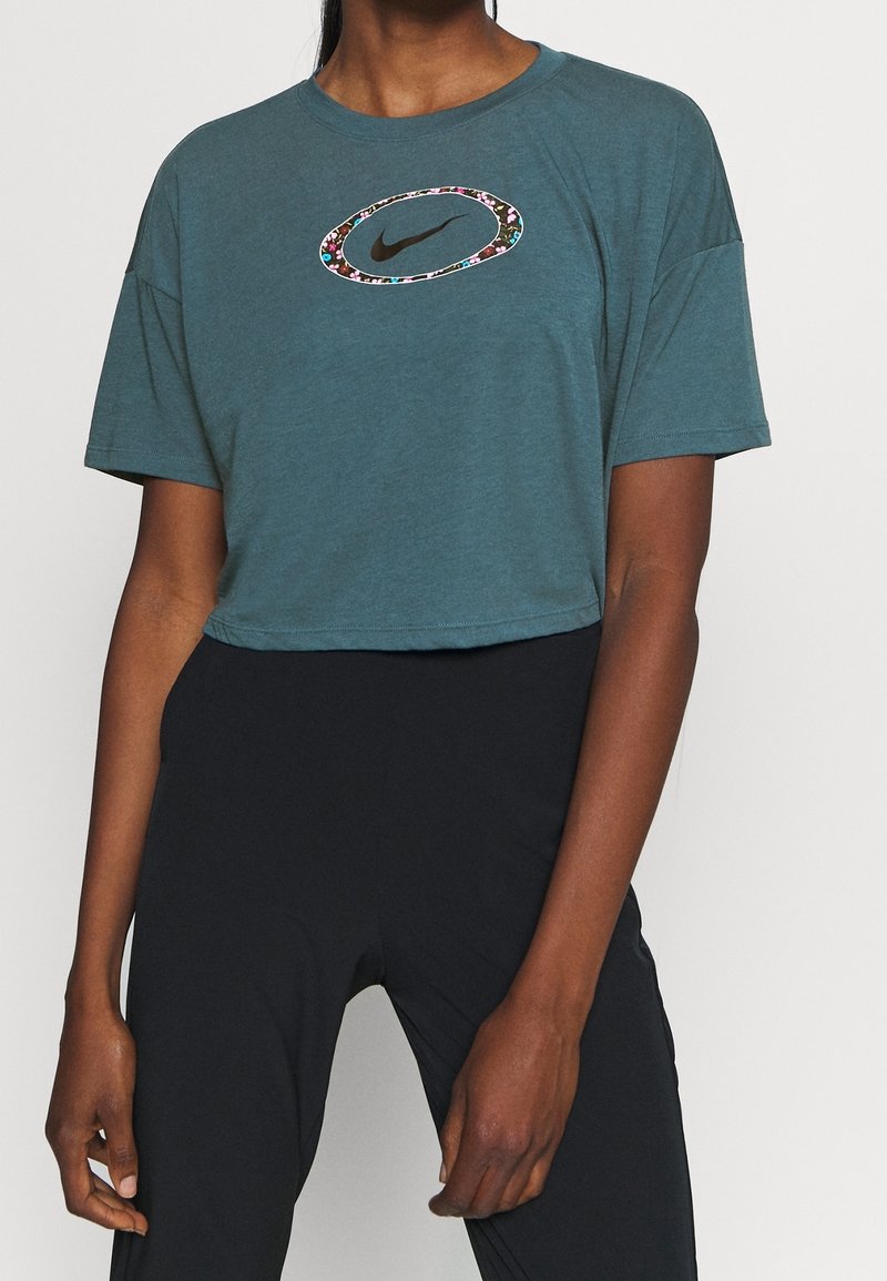 Persona che indossa una maglietta cropped color teal con logo Nike bordato di fiori e leggings neri a vita alta, in piedi su uno sfondo semplice.