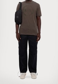 Man wearing brown Armani Exchange T-shirt, black cargo pants, white sneakers, and a black shoulder bag against a plain background.