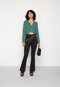 Cardigan verde curto com decote em V e fecho de botão, combinado com calças pretas de cintura alta e boca de sino com um detalhe de corrente. Mala preta.