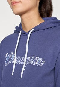 Navy blue hoodie made of soft fabric, featuring white drawstrings and embroidered "Champion" logo in a cursive font on the front.