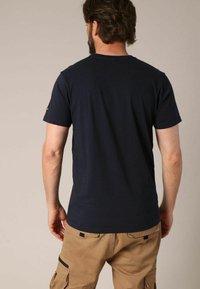 Homme aux cheveux bruns et à la barbe, portant un t-shirt uni bleu foncé à manches courtes et un pantalon cargo beige clair, tourné le dos contre un fond uni.