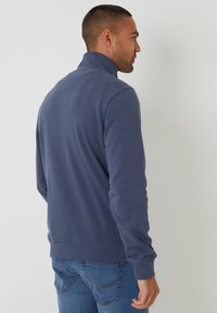 Sudadera de cuello con cremallera en azul marino hecha de una tela suave. Presenta mangas largas y un dobladillo acanalado. Se lleva con jeans azules, mostrando un diseño casual.