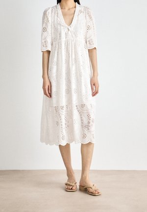 Femme portant une robe midi blanche à motif ajouré, manches ballon, col en V avec lien, et ourlet festonné, associée à des sandales à talons beiges.