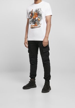 T-shirt bianca in cotone con un design grafico che presenta elementi di basket e testo. Abbinata a pantaloni cargo neri e scarpe sportive nere.