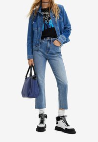 Jeansjacke, grafisches schwarzes T-Shirt, hochtaillierte hellblaue Jeans und schwarz-weiß gestreifte Knöchelstiefel mit Schnürsenkeln; hält eine blaue Tasche.