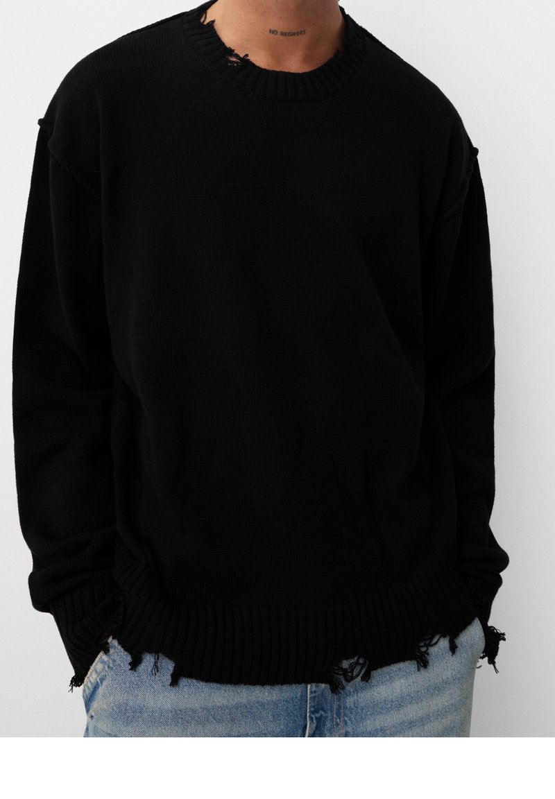 Personne portant un pull en maille noire usé avec des bords effilochés et un jean bleu, les mains dans les poches, cadrée au niveau du cou et de la taille.