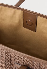Tkaná béžová taška s textúrovaným vonkajškom a hnedými koženými detailmi. Vo vnútri je hladká svetlohnedá podšívka a zapínanie na zatrhávací cvok.