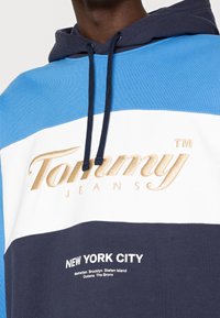 Felpa color block blu, blu navy e bianca con cordini. Presenta il logo dorato "Tommy Jeans" e il testo bianco "NEW YORK CITY" sul davanti.
