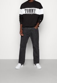 Sudadera de cuello redondo en blanco y negro con el logo "TOMMY JEANS", combinada con vaqueros de corte holgado en gris oscuro y zapatillas blancas con detalles en negro.
