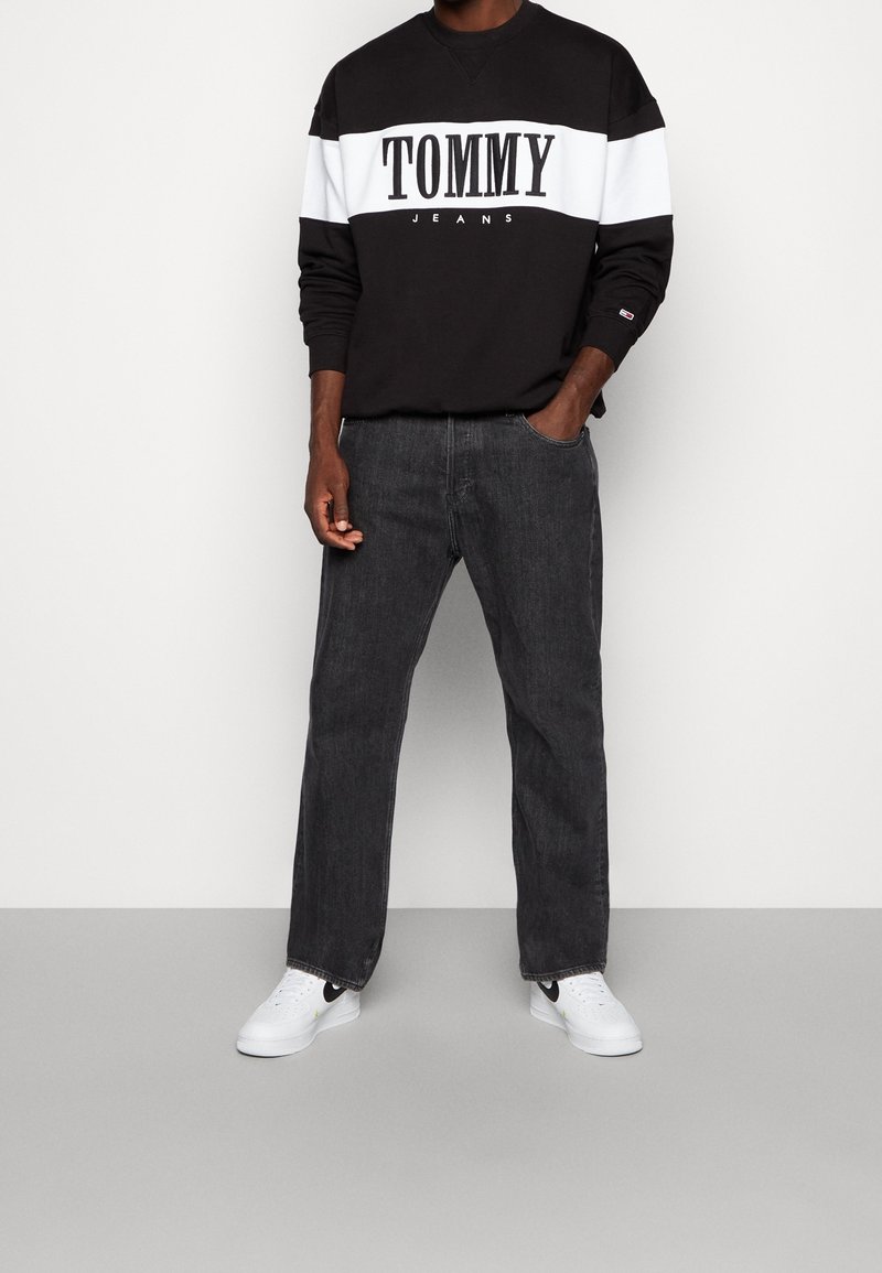Sudadera de cuello redondo en blanco y negro con el logo "TOMMY JEANS", combinada con vaqueros de corte holgado en gris oscuro y zapatillas blancas con detalles en negro.
