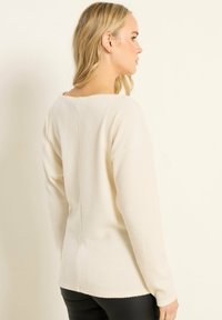 Langärmliger Creme-Pullover mit gerippter Textur, runder Ausschnitt und geradem Rückennaht. Kombiniert mit eng sitzenden schwarzen Hosen.