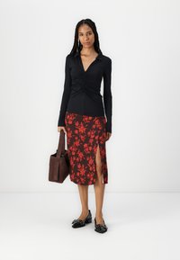 Svart langermet topp med rynkede detaljer, sammen med et rødt skjørt med blomsterdesign og sideløsning. Brun håndveske, svarte flate sko.