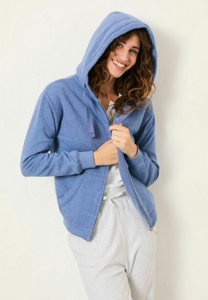 Blue zip-up hoodie made of soft fabric, featuring a large hood, front pockets, and ribbed cuffs, worn over a white top and gray pants.