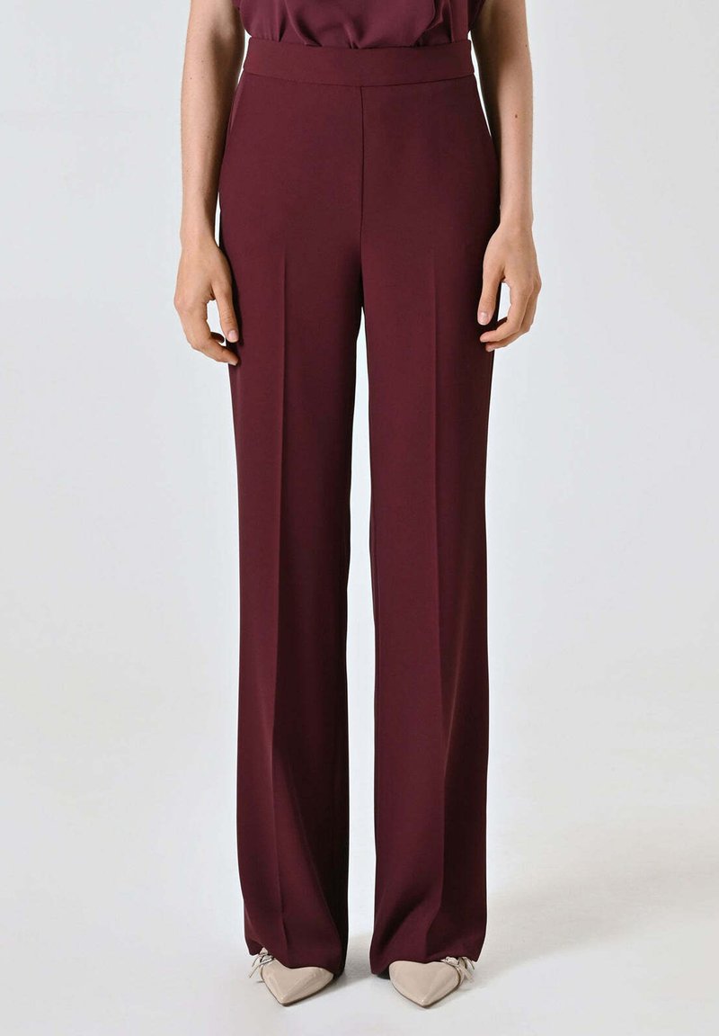 Pantaloni a gamba larga color bordeaux realizzati in tessuto liscio con vita alta e pieghe frontali. Abbinati a scarpe appuntite di colore chiaro.