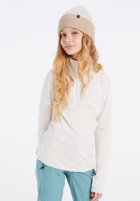 Licht beige langemouwen top met een hoge kraag en een ritssluiting aan de voorkant. Gecombineerd met tealbroeken met verstelbare zijbanden. Wol gebreide beanie.