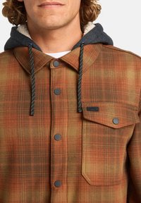 Chaqueta de cuadros con tonos naranjas, marrones y verdes, botones a presión negros y un forro de capucha gris con una textura suave.