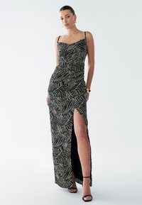 Vestido maxi negro con patrón de hojas beige, silueta ajustada, abertura lateral, tirantes finos y tela suave. Incluye tacones negros con tiras.