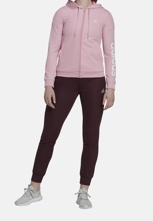 Femme portant un sweat à capuche zippé rose clair avec capuche et un pantalon de jogging Adidas violet foncé, debout les mains détendues le long du corps.