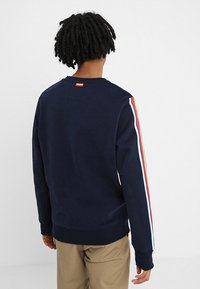 Persona con trenzas cortas vistiendo un sudadera marina con rayas rojas, blancas y amarillas en las mangas, y pantalones beiges, de espaldas.