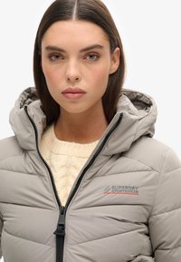 Veste matelassée gris clair avec capuche, dotée d'une fermeture éclair noire, de coutures diagonales et d'un logo subtil en rouge et gris sur la zone gauche de la poitrine.