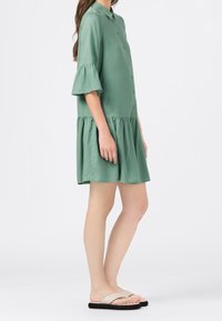 Krótkosłomkowa sukienka z kołnierzykiem w miękkim zielonym materiale z rozkloszowanym, drapowanym dołem. Nosi się ją z beżowymi klapkami.