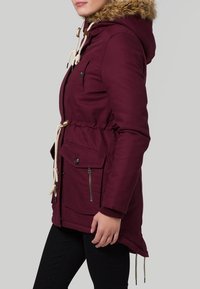 Burgundy parka-jacka med foder i päls, dragsko i midjan, frontdragkedja och klafffickor, samt elastiska ärmslut. Slät tygtextur.