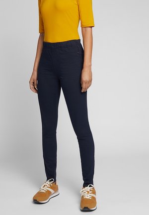 Persoon die strak zittende marineblauwe broek, mosterdgele korte mouw top en mosterdgele sneakers met witte veters draagt, staand tegen een effen achtergrond.