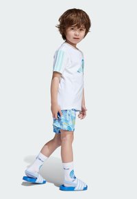 Vit kortärmad skjorta med turkosa detaljer, blåmönstrade badshorts, vita strumpor med blått mönster och blåa sandaletter, designade för barn.