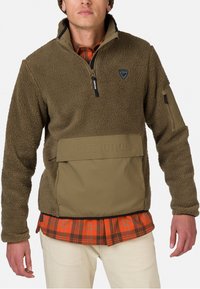 Pullover in pile verde oliva con collo a zip, dettagli neri e tasca frontale con il logo "Rossignol"; indossato sopra una camicia a quadri arancione.