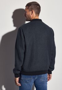 Man in dark gray jacket and blue jeans standing with back to camera casting a shadow on a white wall.
