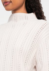 Detailní záběr na ženu oblečenou v krémovém, žebrovaném svetru s vysokým výstřihem a texturovanými svislými vzory.