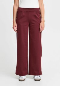 Burgundyröda byxor med vida ben och en mjuk textur, med hög midja och framfållar. Bärs med vita sneakers mot en enkel bakgrund.