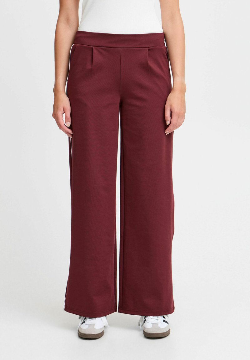Burgundyröda byxor med vida ben och en mjuk textur, med hög midja och framfållar. Bärs med vita sneakers mot en enkel bakgrund.