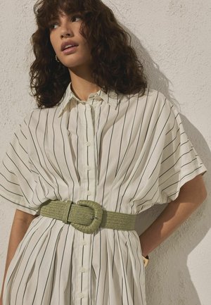 Kvinde med krøllet brunt hår iført en hvid kortærmet kjole med sorte lodrette striber og et bredt grønt bælte, står op ad en struktureret væg.