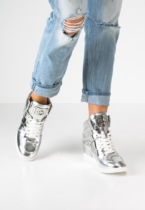 Osoba w jasnoniebieskich podartych dżinsach i błyszczących srebrnych wysokich trampkach z białymi sznurowadłami, stojąca na białej podłodze.