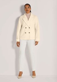 Vrouw in een crèmekleurige dubbelzijdige wollen jas over een beige coltrui, witte slanke broek en beige puntige hakken, staand tegen een effen achtergrond.