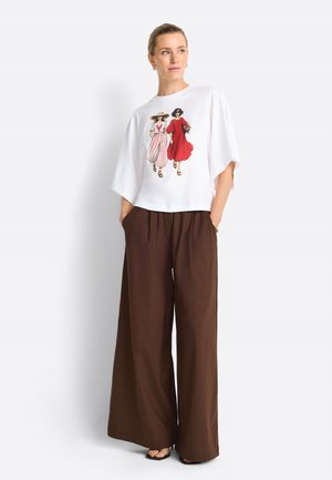 Donna che indossa una maglia bianca oversize con illustrazione di due donne che camminano e pantaloni marroni larghi, in piedi con le mani nelle tasche.