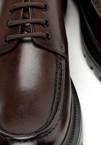 Braune Lederschuhe mit glatter Textur, runder Zehenform, schwarzen Ösen und braunen Schnürsenkeln. Besondere Merkmale sind die detaillierte Naht entlang der Seiten.