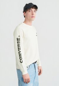 Νέος άνδρας που φοράει λευκό μακρυμάνικο πουκάμισο με την επιγραφή «CONVERSE» στο μανίκι, ανοιχτόγαλανό τζιν, μαύρη ζώνη και μαύρο καπέλο μπέιζμπολ.