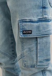 Svetlo modre denim hlače s stranskimi žepi in temno rdečo nalepko "SALTYDOG". Tkanina ima bledijo teksturo in vidno šivanje.