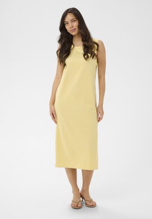 Kobieta o długich ciemnych włosach, ubrana w żółtą sukienkę midi bez rękawów i beżowe sandały na obcasie, stojąca na jednolitym tle.