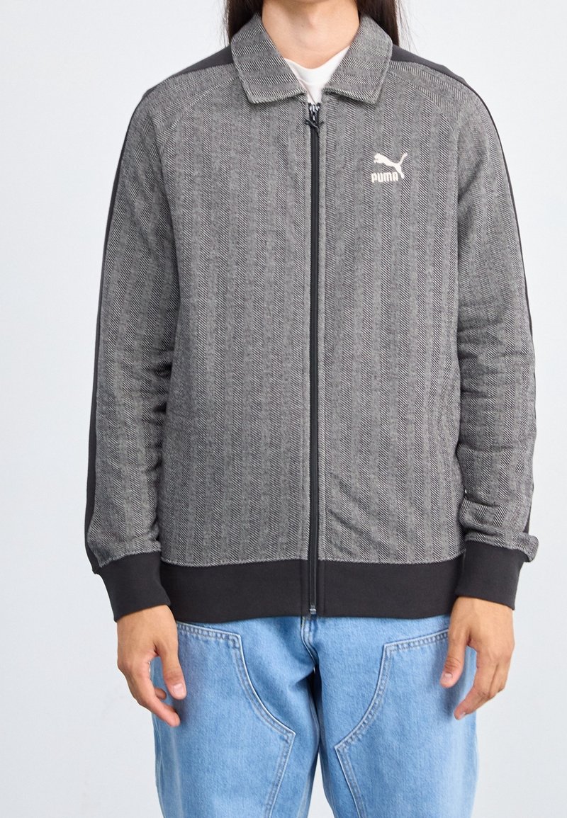 Personne portant une veste zippée grise Puma avec des poignets et un col noirs, associée à un jean bleu clair.