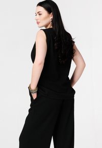 Haut noir sans manches en tissu texturé, associé à un pantalon noir ample. Le mannequin porte un bracelet multi-rangs en accessoire.