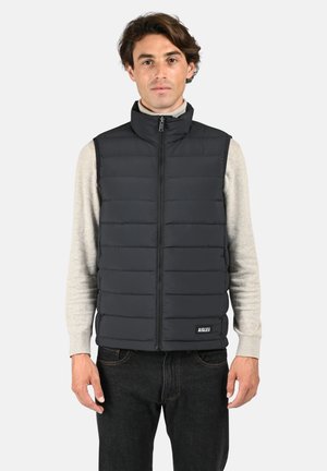 Gilet matelassé noir avec col montant et fermeture éclair à l'avant, porté par-dessus un pull gris clair. Présente une étiquette logo sur le côté inférieur droit.